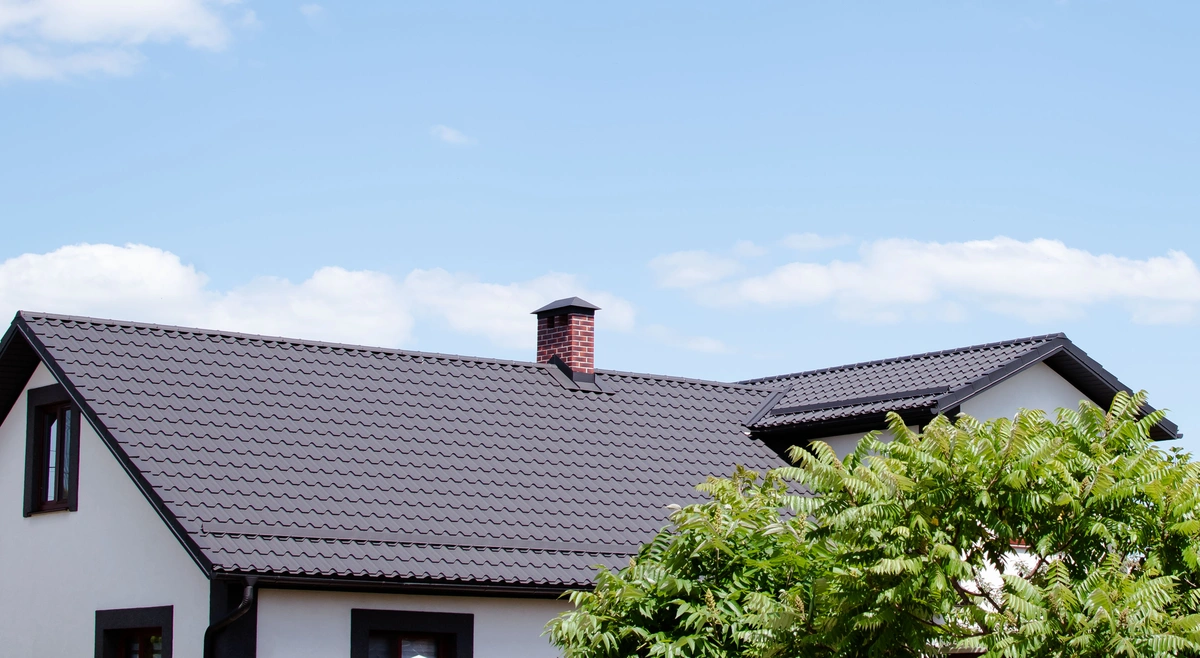 A modern house with a dark gray, metal-tiled roof—one of the popular types of roofs in Florida—and a red brick chimney, partially obscured by green leafy trees under a blue sky with scattered clouds.