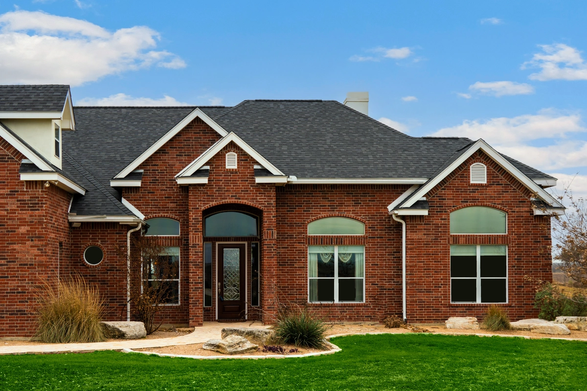 A modern brick house with arched windows and a black shingled roof showcases stylish roof and house color combinations. Surrounded by green grass, rocks, and small landscaped plants, it sits under a partly cloudy blue sky with a glass front door.