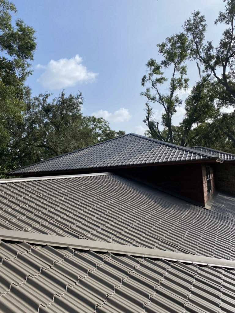 A sloped metal roof with overlapping panels is shown under a sunny sky, surrounded by tall green trees.