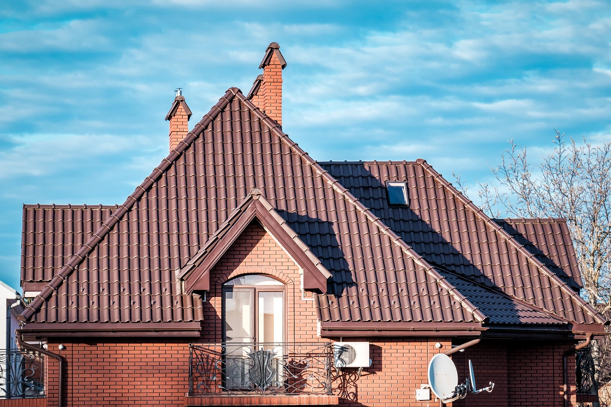 A brick house with a brown metal roof—highlighting the metal roof cost in Florida—two chimneys, a balcony with ornate railings, a satellite dish, an air conditioner, and a small skylight, set against a blue sky with scattered clouds.