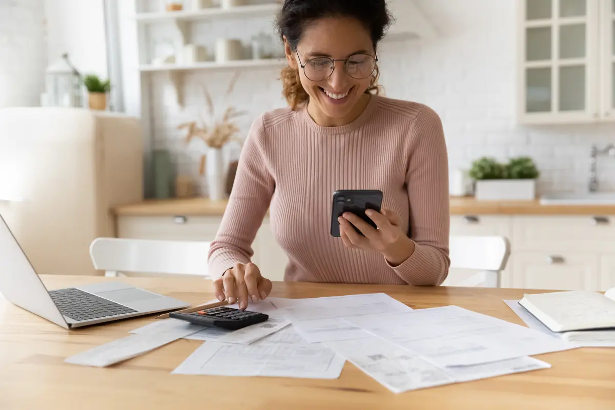 A woman wearing glasses and a pink sweater sits at a table with papers, a laptop, and a calculator. She smiles at her smartphone, possibly checking bills or researching roof inspection cost to manage her finances.