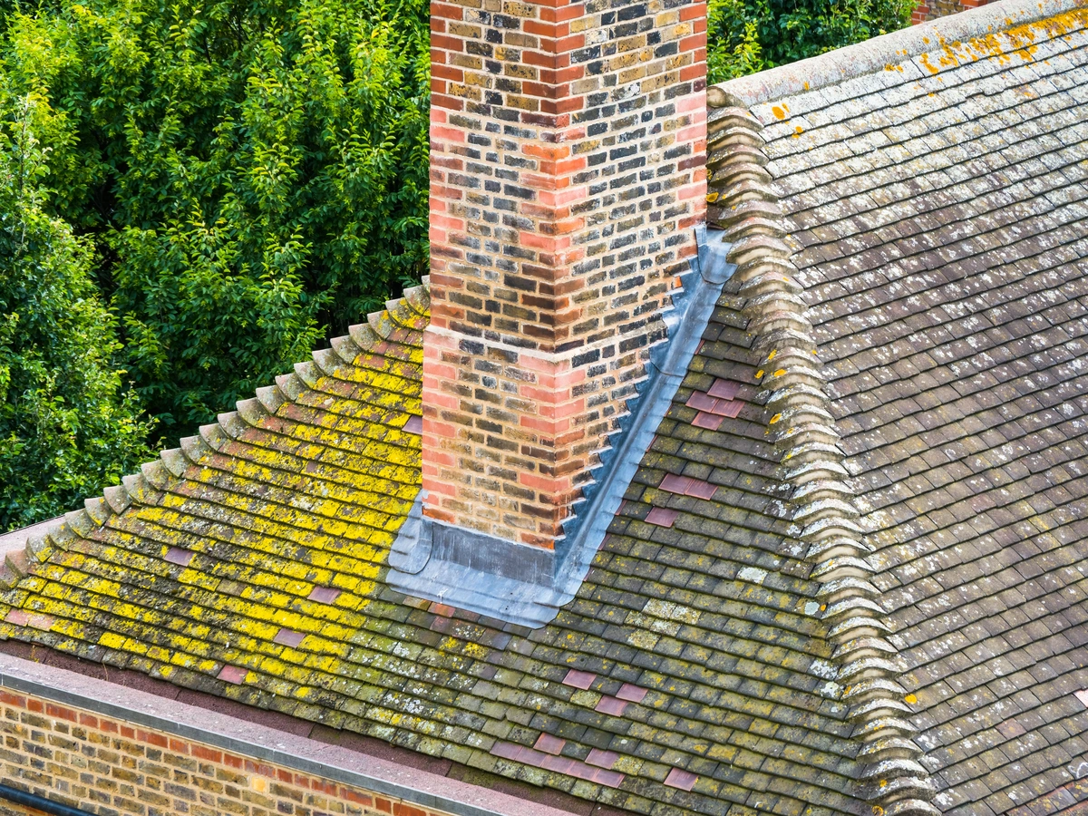 A brick chimney stands on a sloped roof covered with weathered tiles, some with yellow moss or lichen. Roof flashing installation is visible around the chimney’s base. Green trees surround the building in the background.