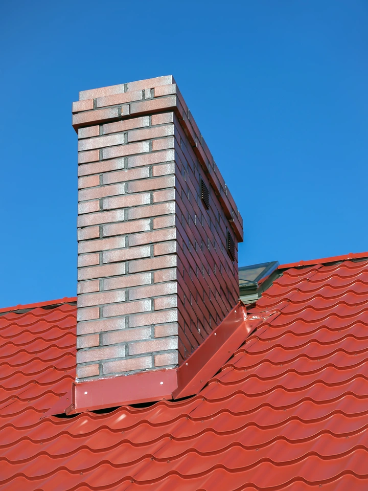 A brick chimney rises from a red tiled roof against a clear blue sky, with sunlight highlighting the chimney’s texture, the roof’s wavy pattern, and the neat lines of roof flashing where they meet.