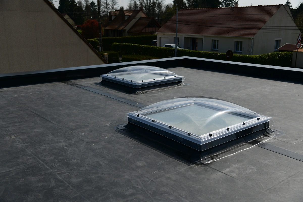 Two rectangular skylights with clear domes are installed on a flat, dark-colored roof, highlighting the differences between EPDM vs TPO roofing materials. Houses with sloped roofs and a few trees are visible in the background.