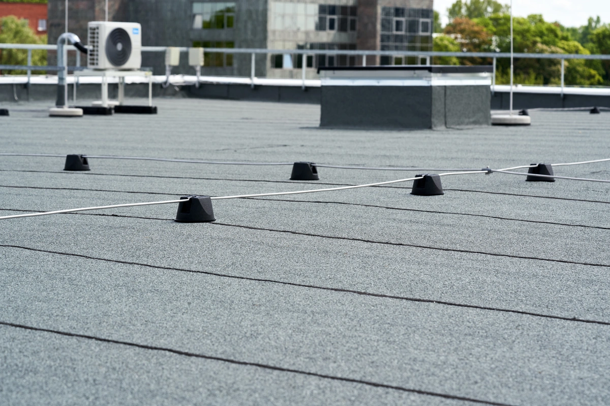 A flat commercial rooftop with black rubber cones supporting a safety cable system, featuring HVAC equipment and buildings in the background—ideal for inspection or flat roof repair needs.