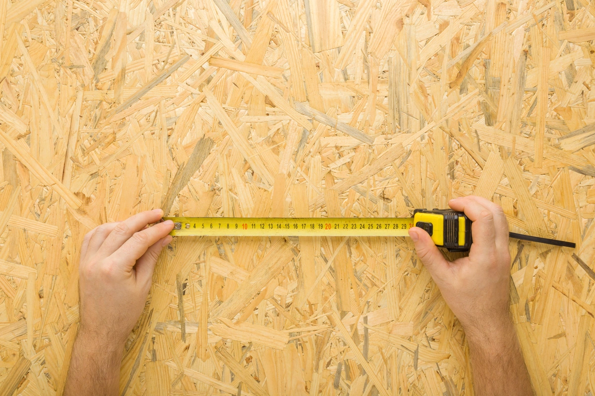 A person holds a yellow tape measure stretched across a sheet of oriented strand board (OSB) wood, measuring its length in centimeters—an essential step in learning how to measure a roof accurately.