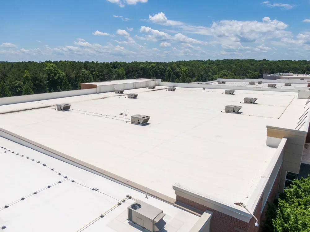 A large, flat commercial building roof with several HVAC units, surrounded by trees under a blue sky with scattered clouds.
