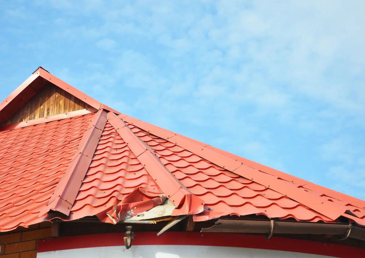 A red metal roof with visible damage and bent panels near the edge, set against a blue sky with some clouds—an example of common metal roof installation mistakes.