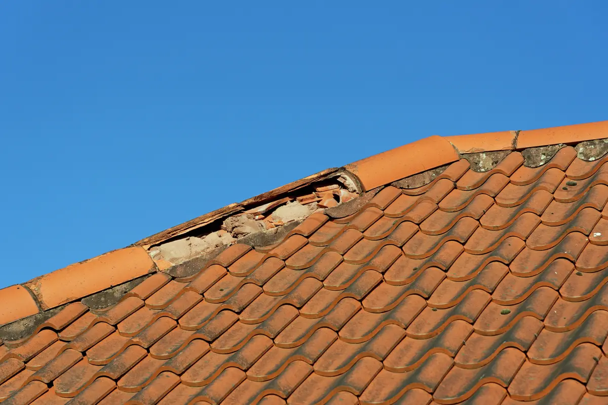 Close-up of a terracotta tiled roof with several broken and missing tiles near the ridge, exposing the underlying structure—a clear sign that tile roof replacement may be needed—set against a clear blue sky.