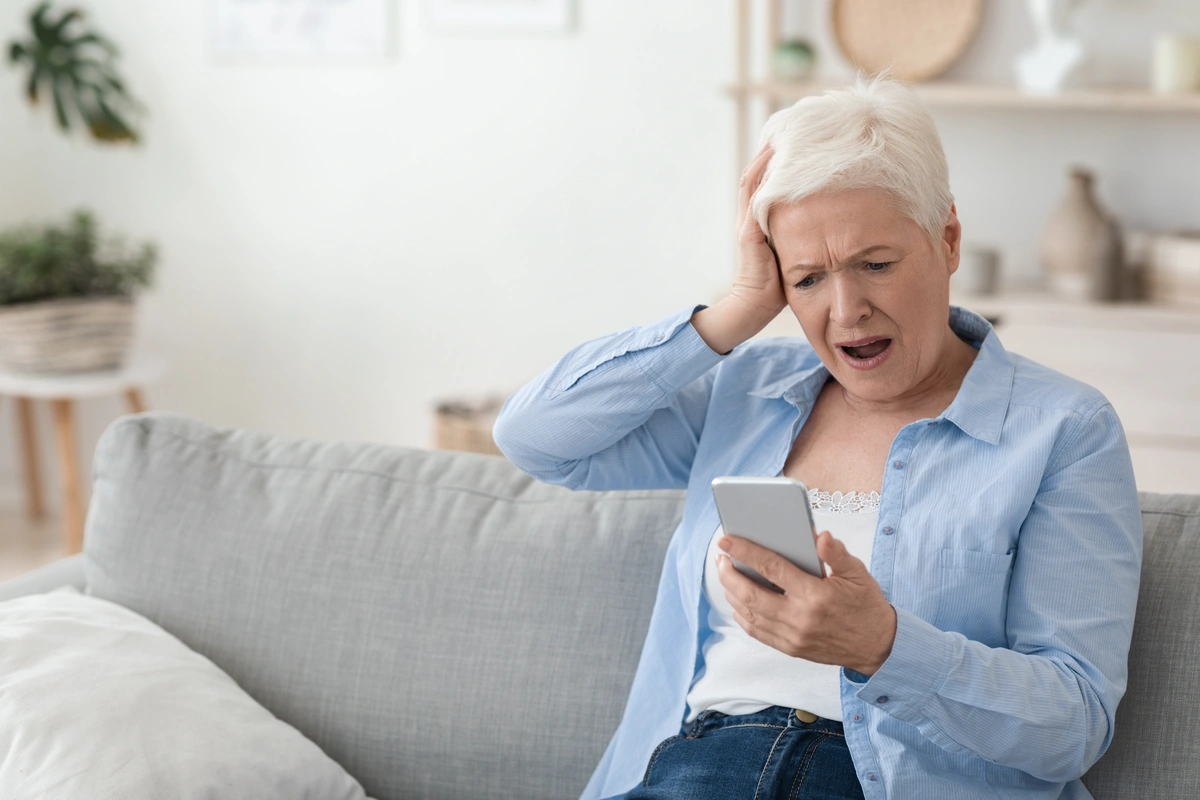 An older woman with short gray hair looks shocked and upset while sitting on a couch, holding her smartphone after reading about florida roofing scams, her other hand pressed anxiously to her head.
