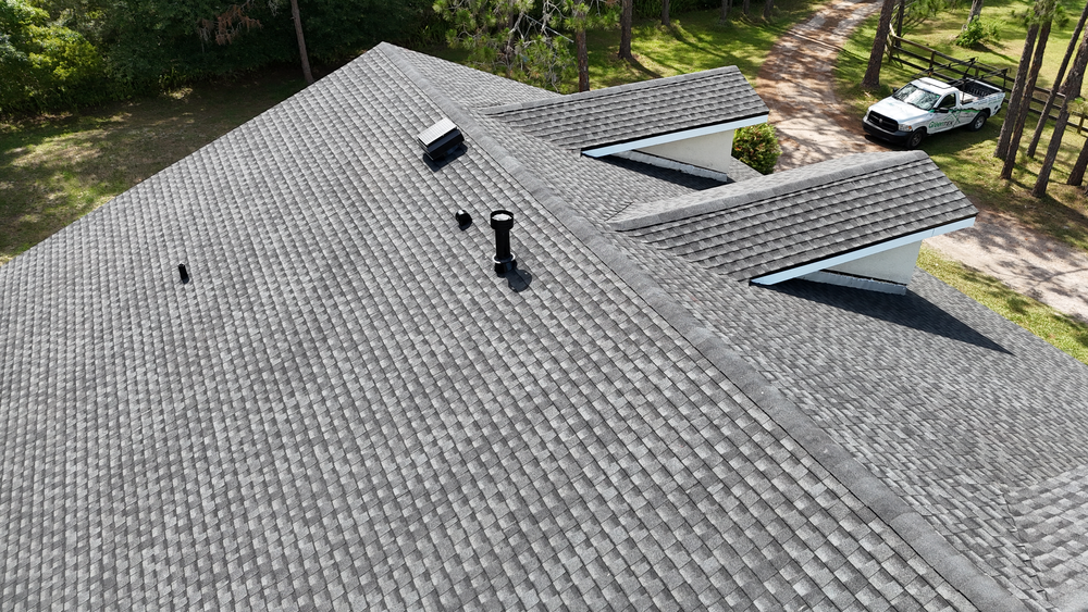 Aerial view of a gray shingled house roof with several vents and a chimney, surrounded by trees and a driveway with a parked pickup truck in the background.