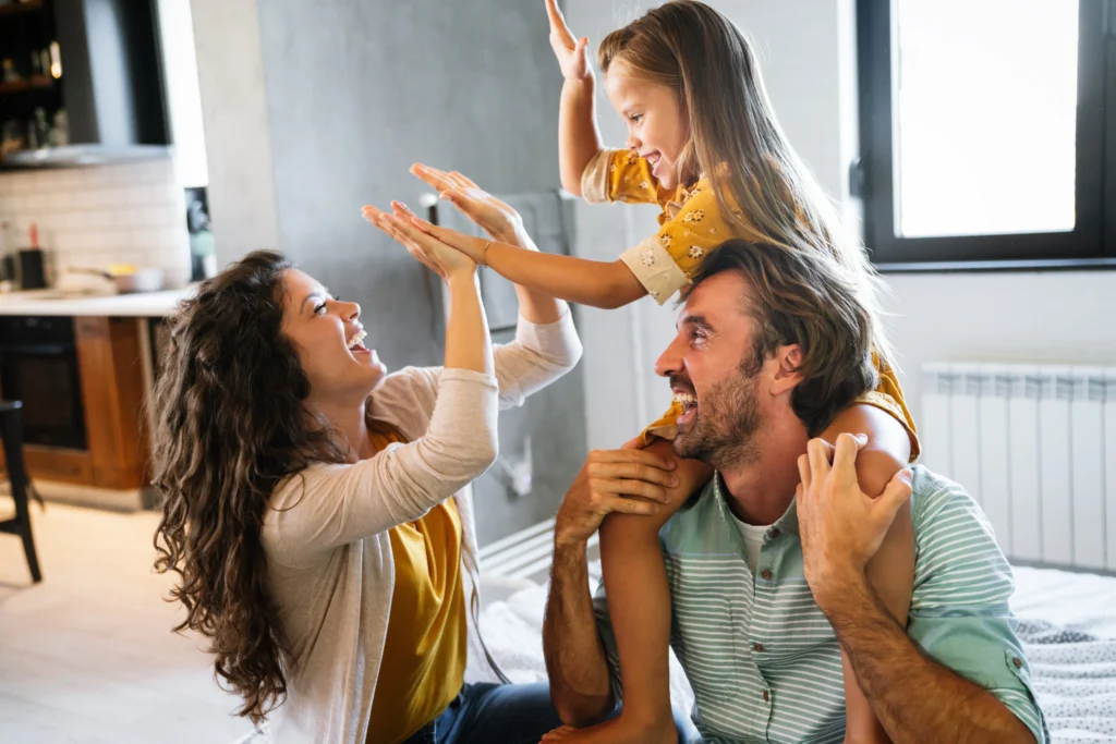 A smiling family enjoys time together at home; a woman and a young girl sitting on a man's shoulders are giving each other high-fives in their bright, modern kitchen-living area, thanks to secure roofing Lakeland residents can trust.