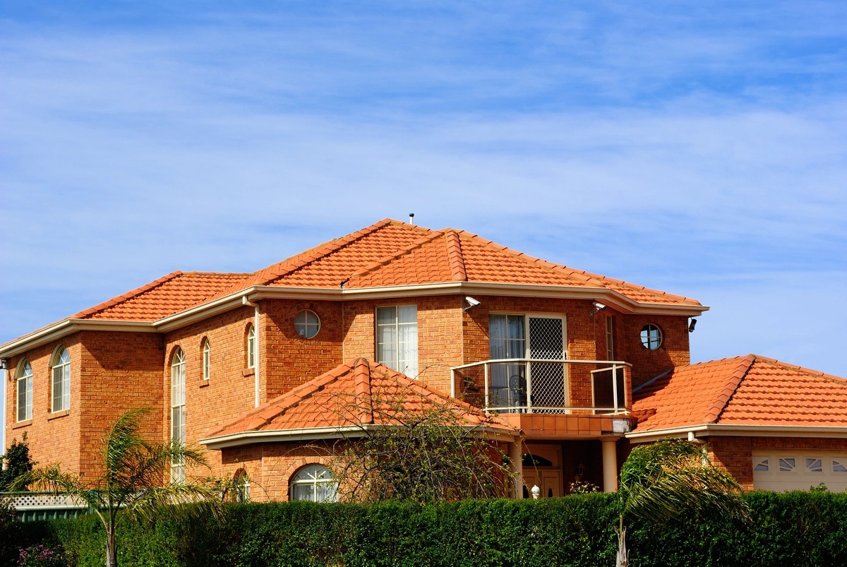 Two-story brick house with orange-tiled roof, balcony, arched windows, and attached garage—recently maintained with expert tile roof repair—surrounded by greenery under a blue sky with wispy clouds.