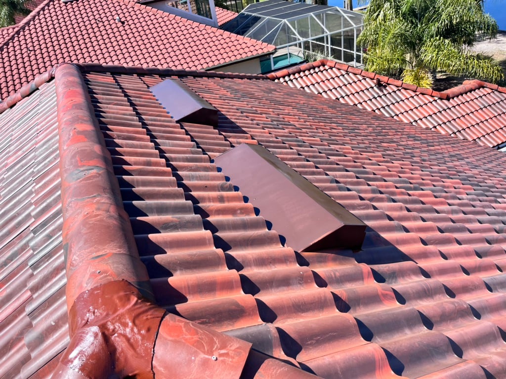 Red tiled roof with two large rectangular metal vents installed on the sloped surface. Nearby rooftops with similar tiles and palm trees are visible in the background.