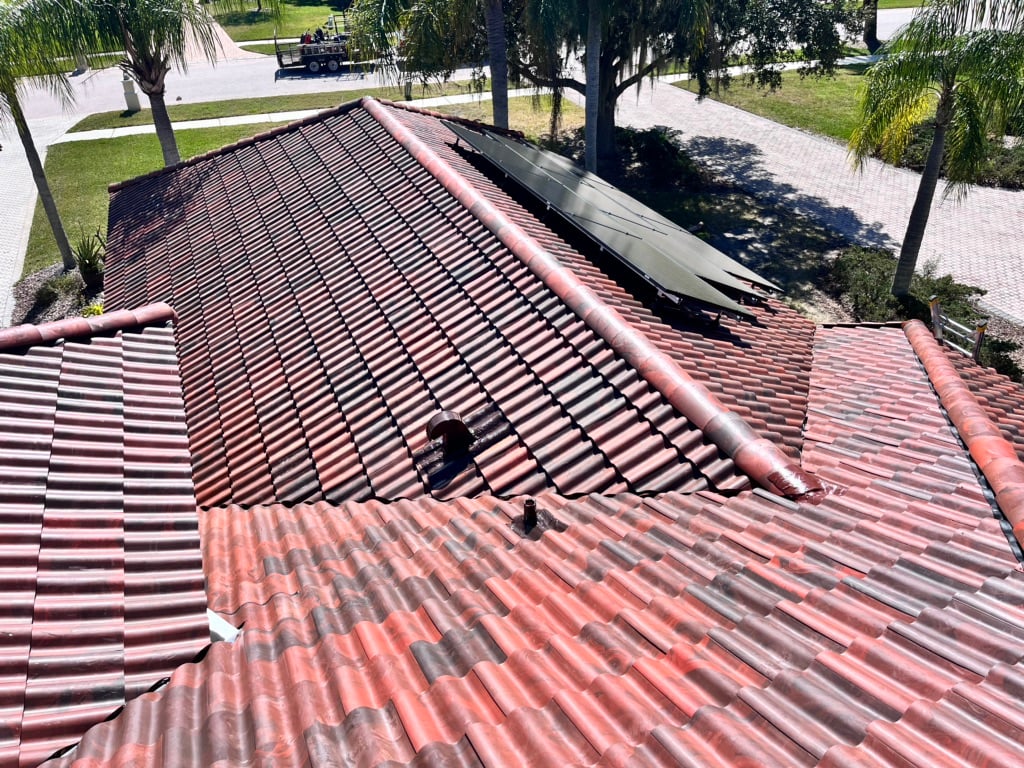 A person in dark clothing works on a red tiled roof near solar panels on a sunny day, surrounded by palm trees and paved pathways.