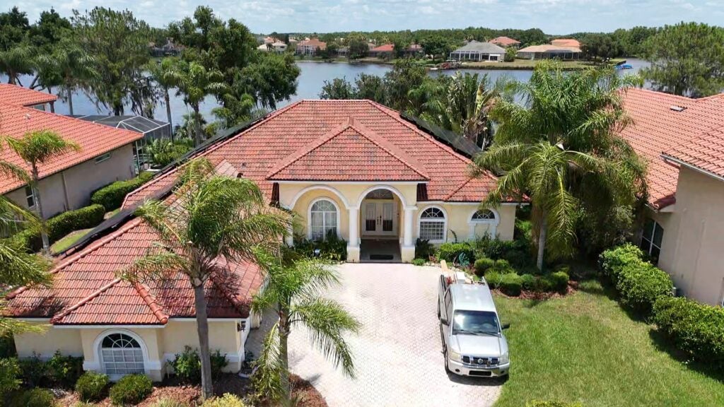 Aerial view of a single-story house with a red tile roof, cream walls, and an arched entryway, surrounded by palm trees and landscaping. A silver car is parked in the driveway. A lake and other houses are visible in the background.