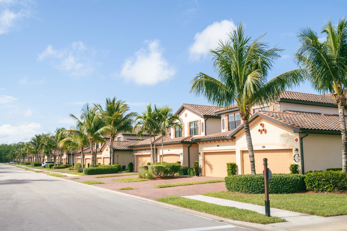 A row of modern, beige townhouses with red-tiled roofs and palm trees lining the street under a blue sky with scattered clouds. Manicured lawns and driveways reflect a clean, suburban neighborhood built to withstand the Florida heat.