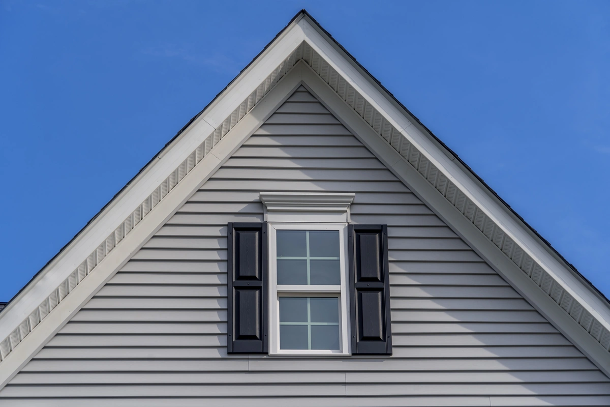 The image shows the upper portion of a house with light gray siding, a single window with black shutters, and a sharply peaked gable roof—an example often compared in discussions of hip roof vs gable roof—set against a clear blue sky.