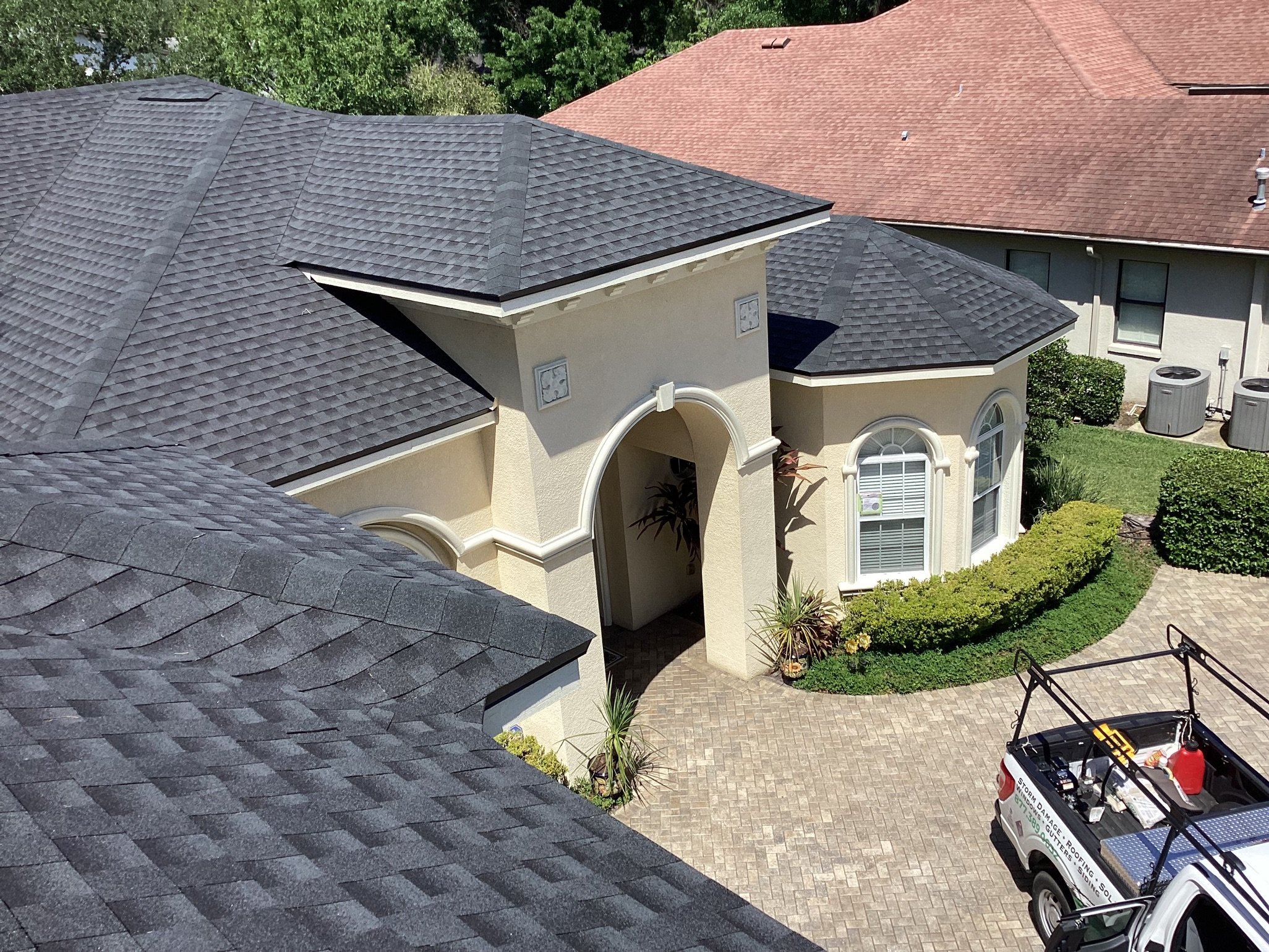 A view from above of a beige house with arched entryway, dark gray shingle roof, brick driveway, manicured bushes, and a white work truck with a roof rack parked outside. Neighboring houses are visible.