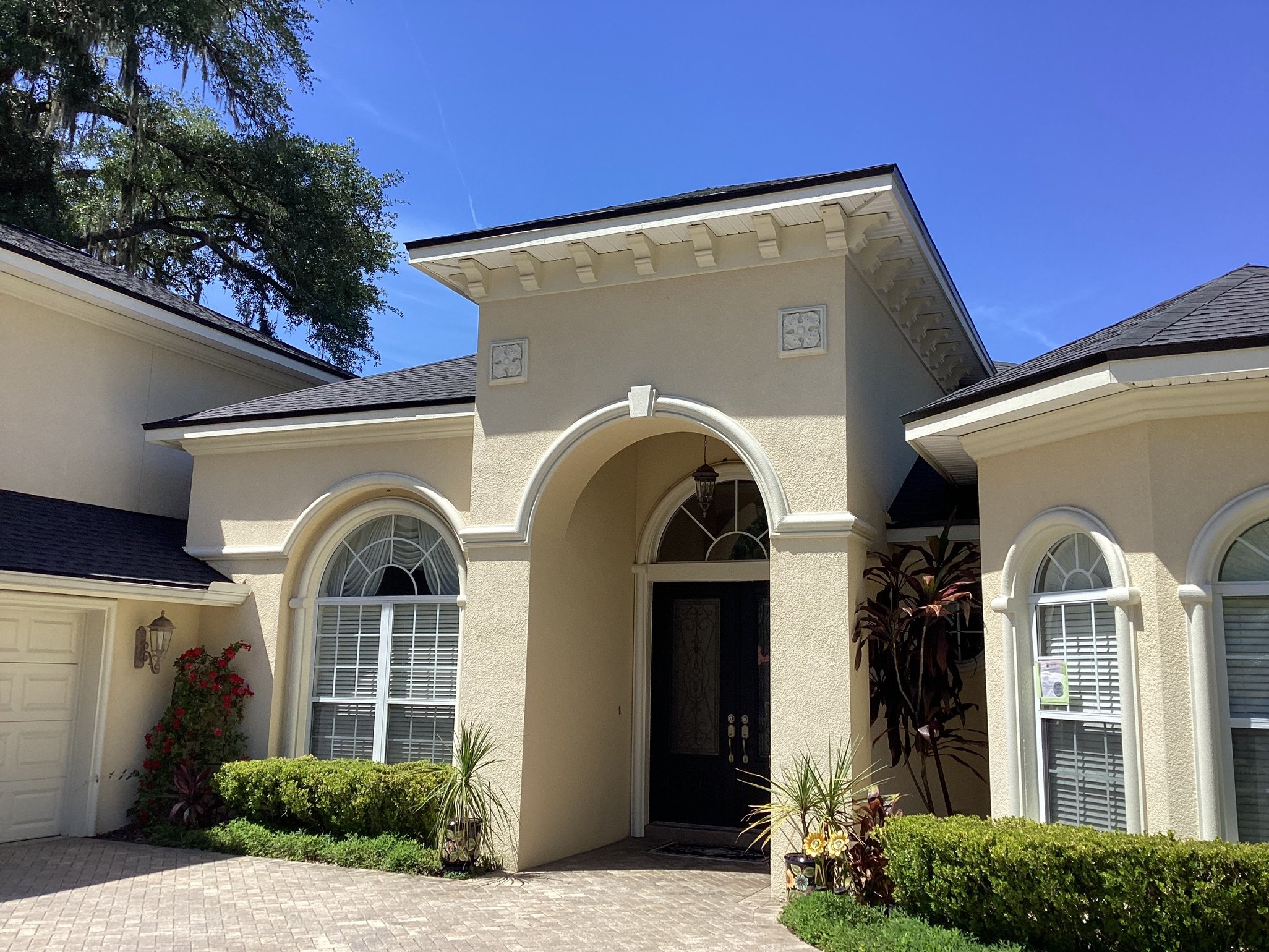 Beige, stucco house with arched windows and a double-door entry under a large archway, surrounded by green shrubs and tropical plants, under a clear blue sky.