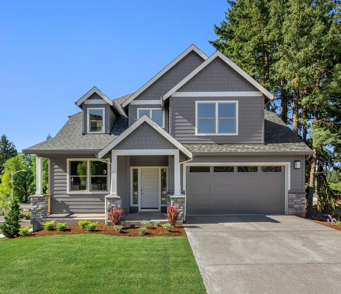 A modern two-story gray house with a two-car garage, front porch, manicured lawn, and young landscaping sits among tall evergreens under a clear sky—a great place to learn how to pay for a new roof when updating your home.