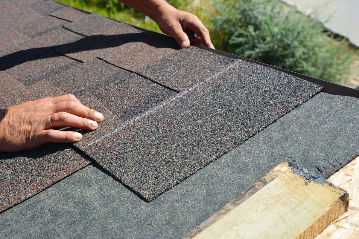 Close-up of hands demonstrating how to install roof shingles, carefully aligning asphalt shingles over underlayment near the roof edge on a sunny day.