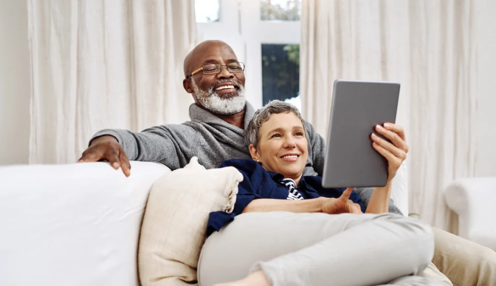 An older couple sits together on a couch, smiling and relaxed. The woman is holding a tablet, and both appear to be enjoying browsing roofing Brandon options on the screen. Light curtains and a window are in the background.