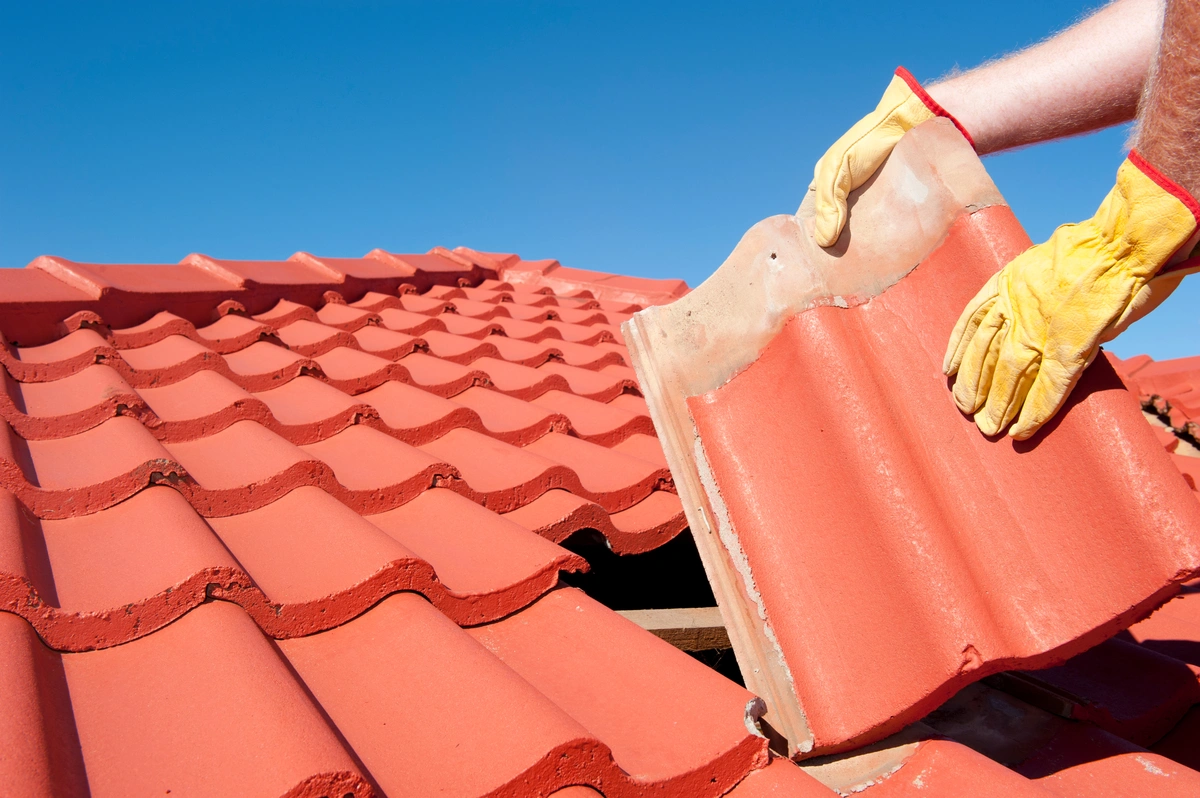 A person wearing yellow work gloves is replacing a red clay roof tile on a house under a clear blue sky, carefully lifting one tile to fit into place among the rows—an essential task when considering tile roof cost and maintenance.