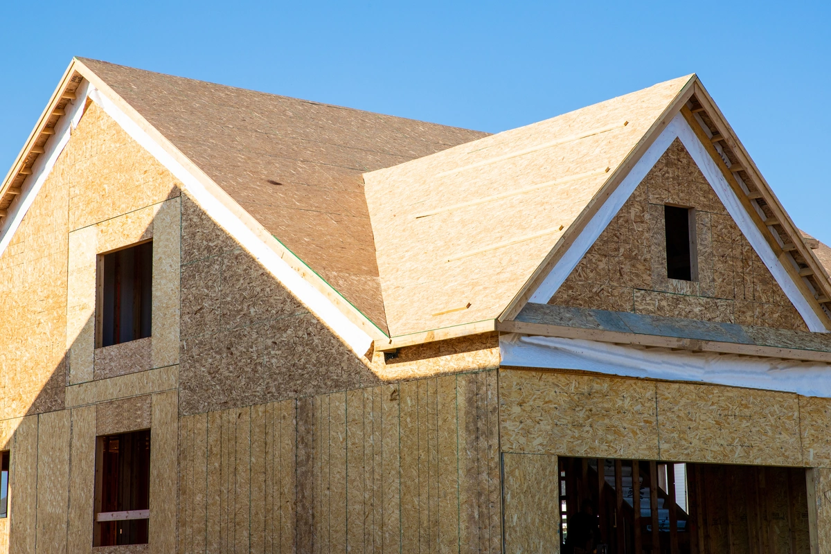 A close-up view of a house under construction, showing wooden framing and OSB sheathing on the exterior walls and redecking a roof, set against a clear blue sky.