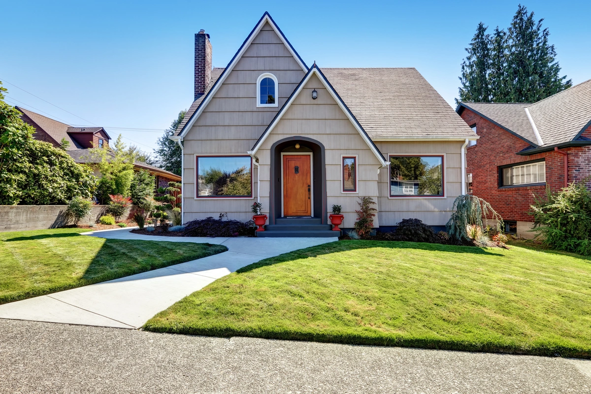 A charming beige house with a steep roof boasting a roofing warranty, central wooden door, and large front windows sits on a neatly manicured lawn with a curved concrete walkway, surrounded by shrubs and potted plants.