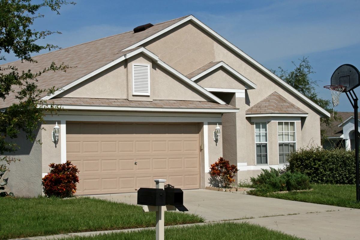 A beige suburban house with a two-car garage, a driveway, green lawn, and basketball hoop sits under a clear blue sky. The front yard features shrubs and small trees—a great place to consider metal roof vs shingles in Florida.