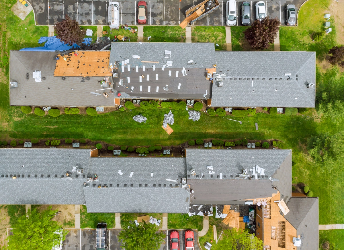 Aerial view of two long apartment buildings with workers repairing roofs, demonstrating how to repair roof shingles blown off. Roofing materials and debris are scattered across rooftops and grass, with parked cars visible near the buildings.