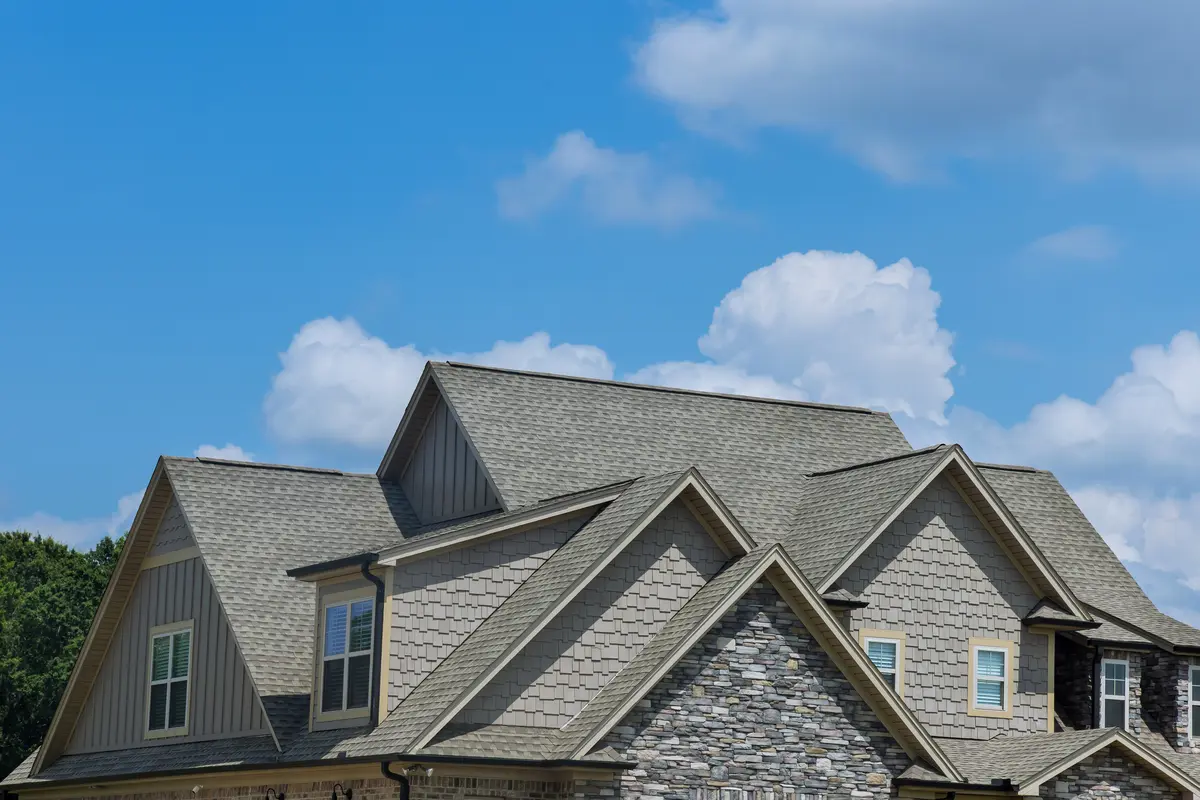 A close-up view of a house roof with multiple gables and peaks, featuring durable class 4 shingles and partial stone siding, set against a blue sky with scattered clouds.