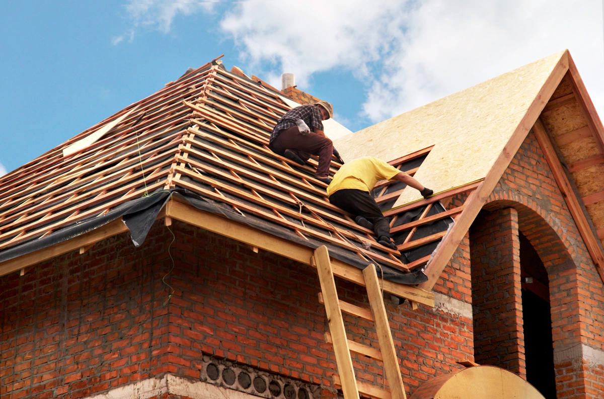 Two workers in casual clothes install wooden planks on the roof frame of an unfinished brick house under a partly cloudy blue sky, carefully avoiding common roofing mistakes. A wooden ladder leans against the roof.