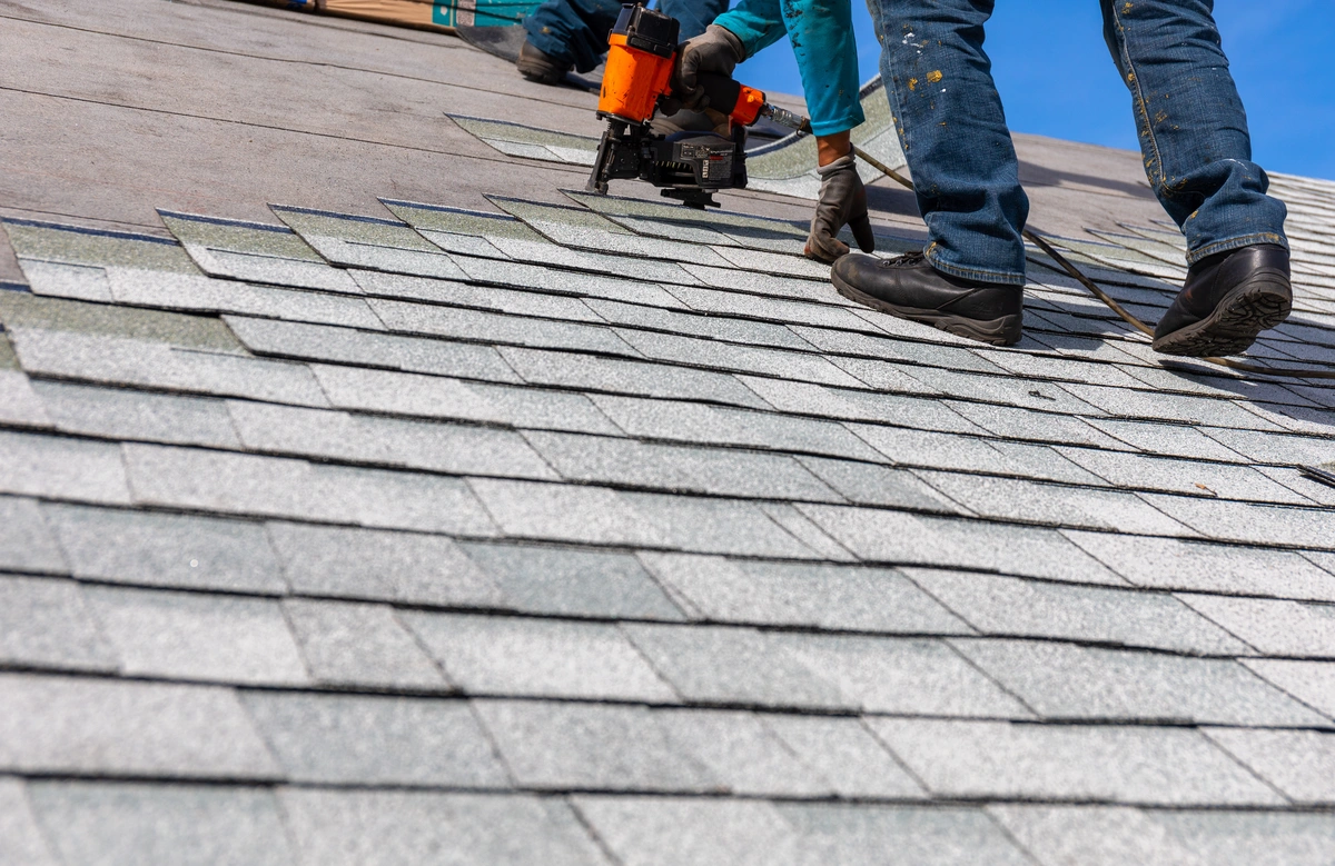 Two workers in jeans and gloves are installing roof shingles with a nail gun on a sloped roof beneath a clear blue sky.