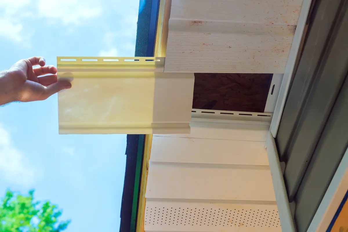 A person installs a piece of light-colored vinyl siding onto the exterior wall of a house, fitting it into place near the roofline under the eaves, alongside a soffit vent, on a sunny day.