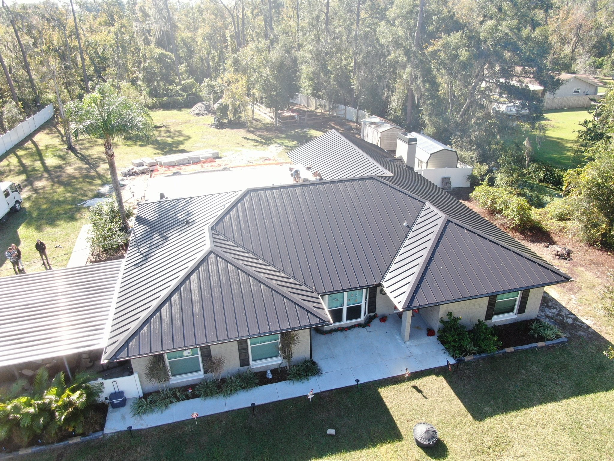 Aerial view of a single-story house with a dark metal roof, surrounded by grass, trees, and a driveway. The backyard contains a concrete slab and several sheds. People and vehicles are visible on the property.