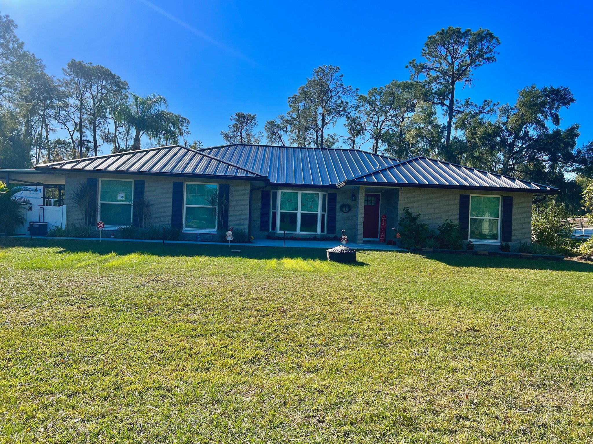 A single-story house with a metal roof, three sets of windows in front, a large grassy lawn, and trees in the background under a clear blue sky.