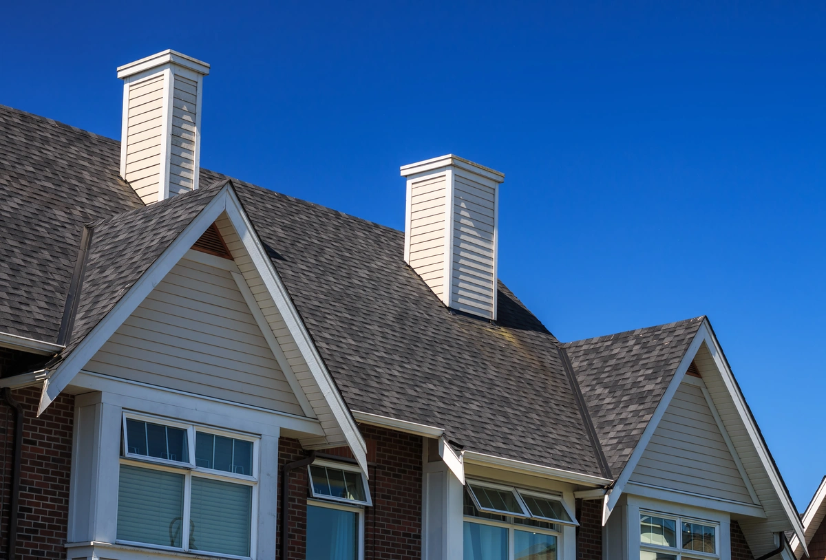 A close-up view of a house roof with dark gray shingles and a roof overlay, featuring two white chimneys and several gabled windows, set against a clear blue sky.
