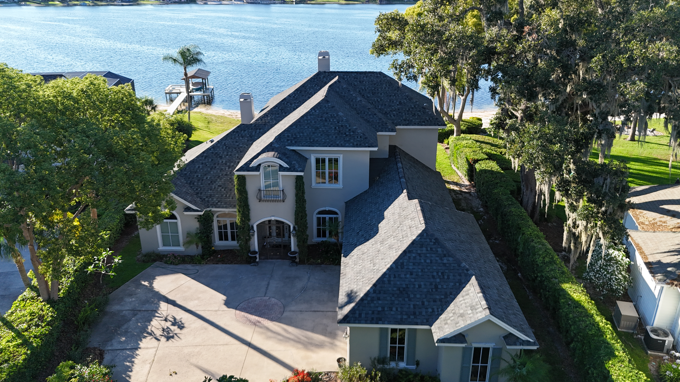A two-story house with a gray roof, surrounded by trees and greenery, sits beside a lake with a dock visible in the background on a sunny day.