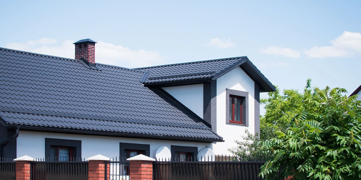 A modern house with white walls, dark trim, and a dark tiled roof—expertly crafted by roofing companies Wesley Chapel FL—featuring a chimney, surrounded by trees and a metal fence with brick pillars under a clear blue sky.