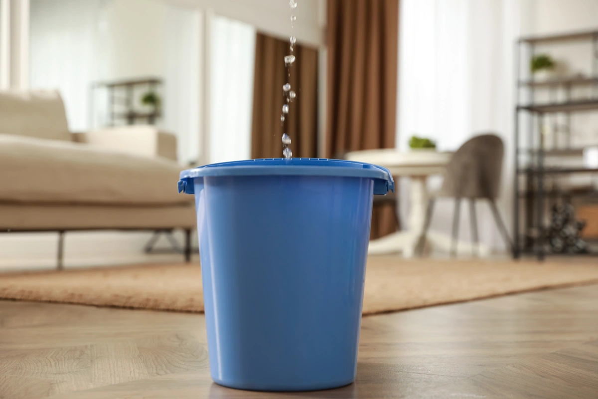 A blue plastic bucket catching water leaking from ceiling, placed on a wooden floor in a modern living room with beige furniture, a rug, and shelves in the background.