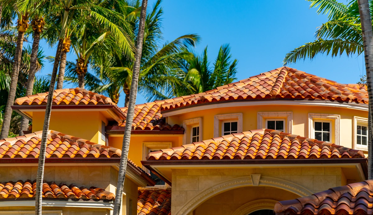 A luxury house with orange terracotta roof tiles and cream-colored walls, expertly crafted by roofing contractors Tampa, surrounded by tall palm trees under a bright blue sky.
