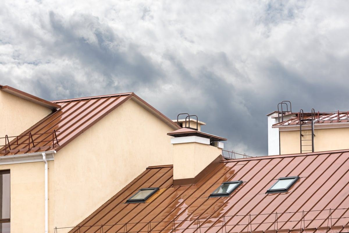 Sloped roofs with brown metal sheet colors, cream-colored walls, skylight windows, and chimneys under a cloudy sky. Ladders and safety railings are visible along the roof edges.