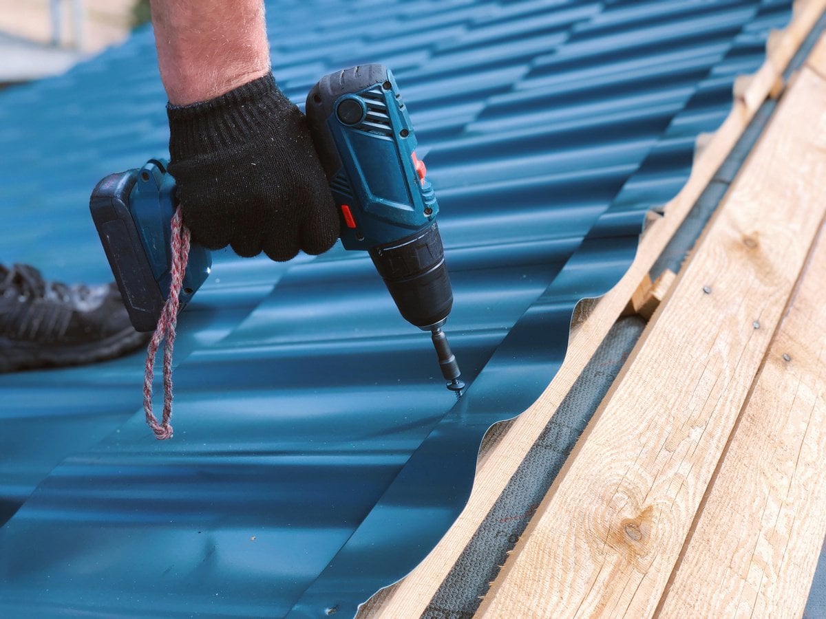 A person wearing a black glove uses a power drill to secure a blue metal roofing sheet onto a wooden roof frame, installing a metal roof over shingles.