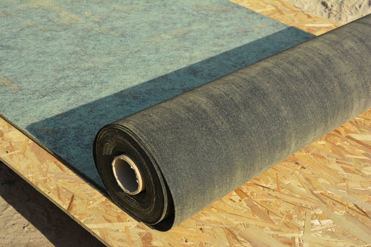 A roll of black roofing felt is partially unrolled on top of wooden boards, ready to be installed as a metal roof underlayment for a roofing project.