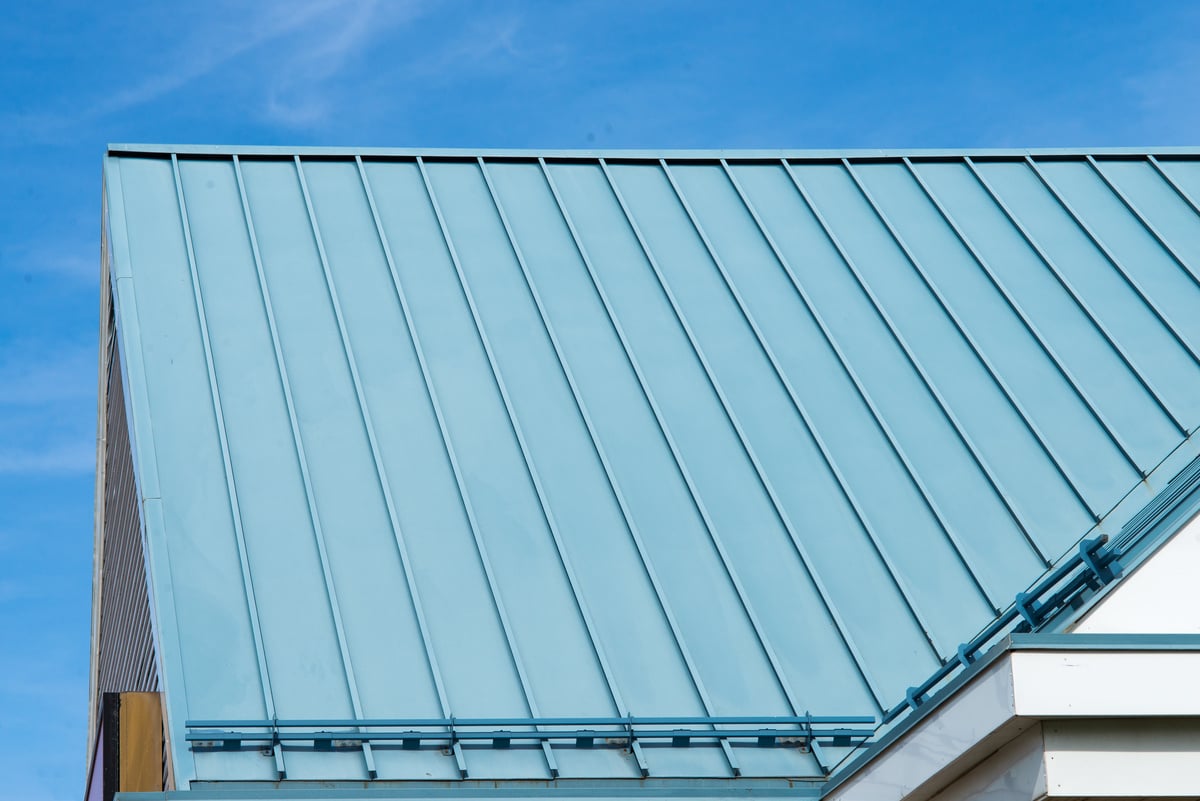 A close-up view of a modern building’s blue metal roof with vertical seams, set against a clear blue sky, highlights the sleek design and durability often considered when comparing metal roof vs shingles cost.