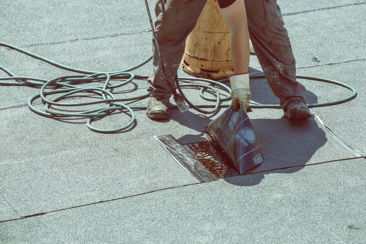 Person in work clothes installing a sheet of modified bitumen roof material with adhesive on a flat roof, holding a roll in one hand and unrolling it onto black sealant, with a hose and equipment nearby.