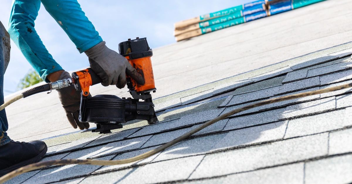 A worker wearing gloves uses a nail gun to install asphalt shingles on a roof under a clear sky, with roofing materials visible in the background—showing part of the process when asking, "How many shingles do I need?.