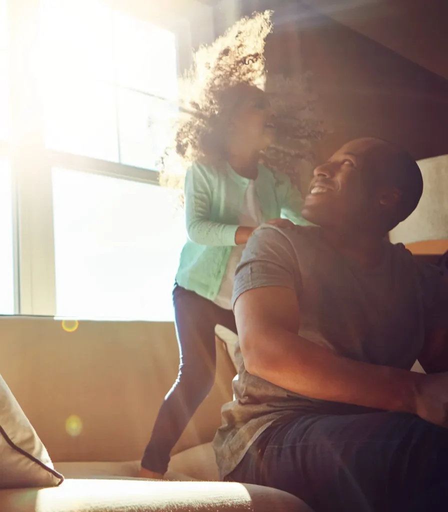 A young girl with curly hair and a big smile plays energetically behind a smiling man sitting on a couch, with bright sunlight streaming through the window behind them.